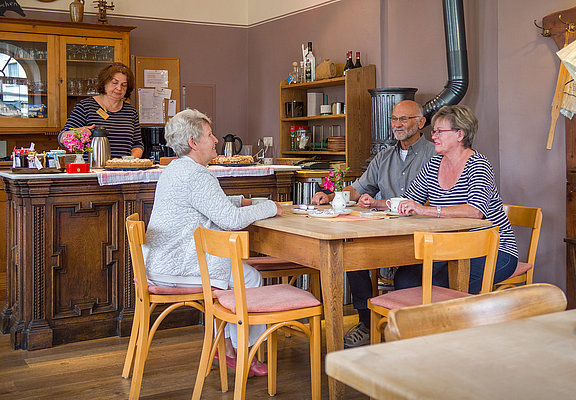 Das Bild zeigt die Gaststube des Hunsrücker Holzmuseums. Drei Personen sitzen am Tisch und trinken Kaffee. Eine Person steht hinter der Theke, auf der auch Kuchen steht.